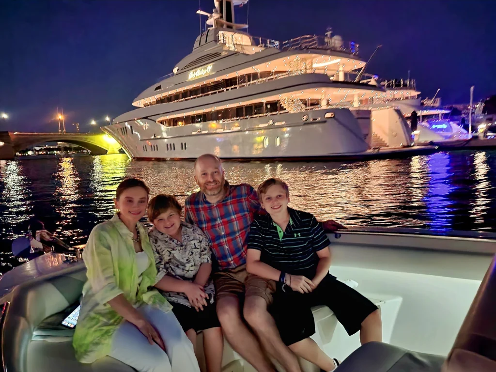 A family with kids posing for a night shot during a sunset cruise with a lit up yacht in the background of West Palm Beach