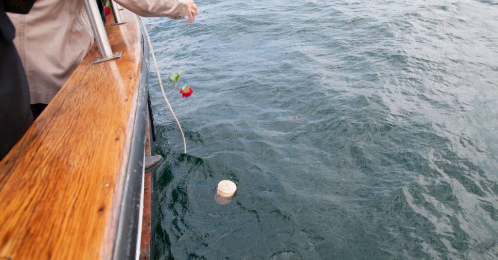 A person wearing a tan jacket leaning over the railing of a boat to drop a red flower into the ocean.