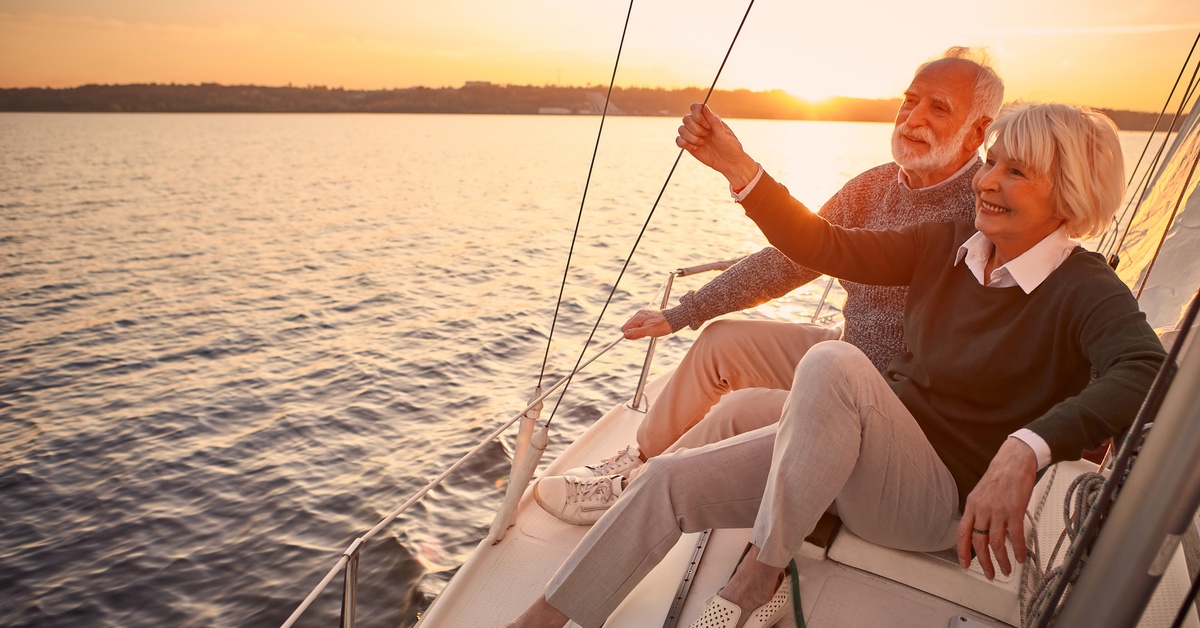 A happy senior couple is relaxing next to each other on the side of a yacht deck, floating in the sea at sunset.