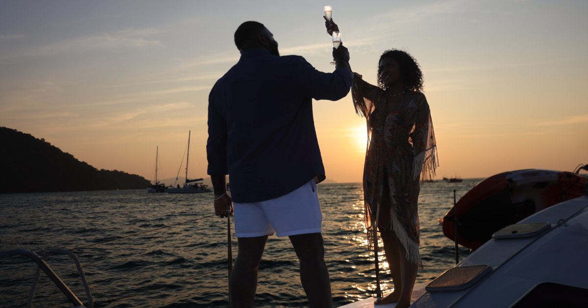 A silhouette of a couple drinking champagne on a private yacht. They're toasting to one another as the sun sets.