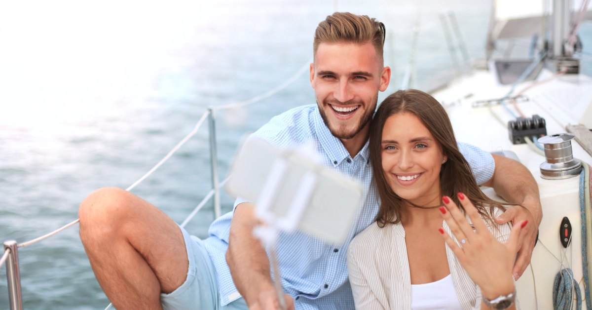 A couple is showing off their recent engagement on a private boat. The woman is using a selfie stick to take a picture with her phone.