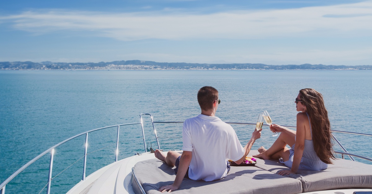 A man and woman clink champagne glasses while seated on gray cushions at the bow of a boat with land in the distance.